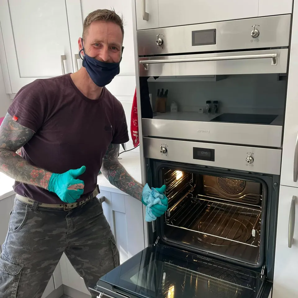 A smiling oven cleaner in a face mask and gloves giving a thumbs up next to a freshly cleaned built-in oven